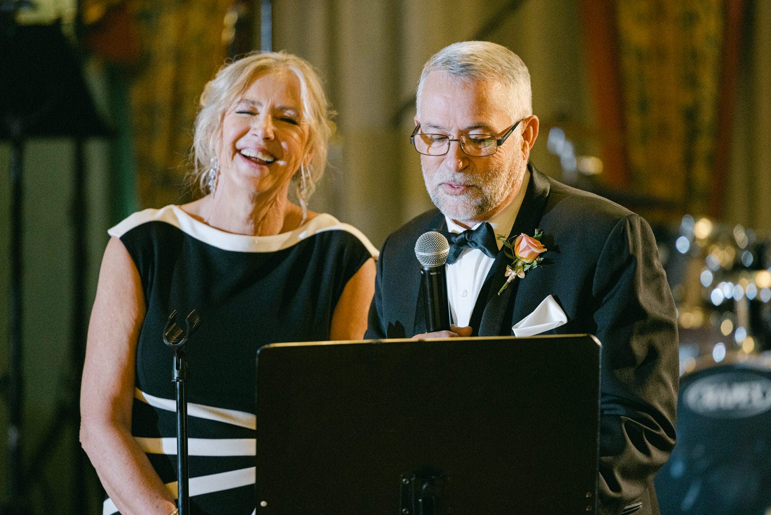 A man and woman are sharing a lighthearted moment during a speech at a wedding reception, with the woman smiling and the man reading from a podium.