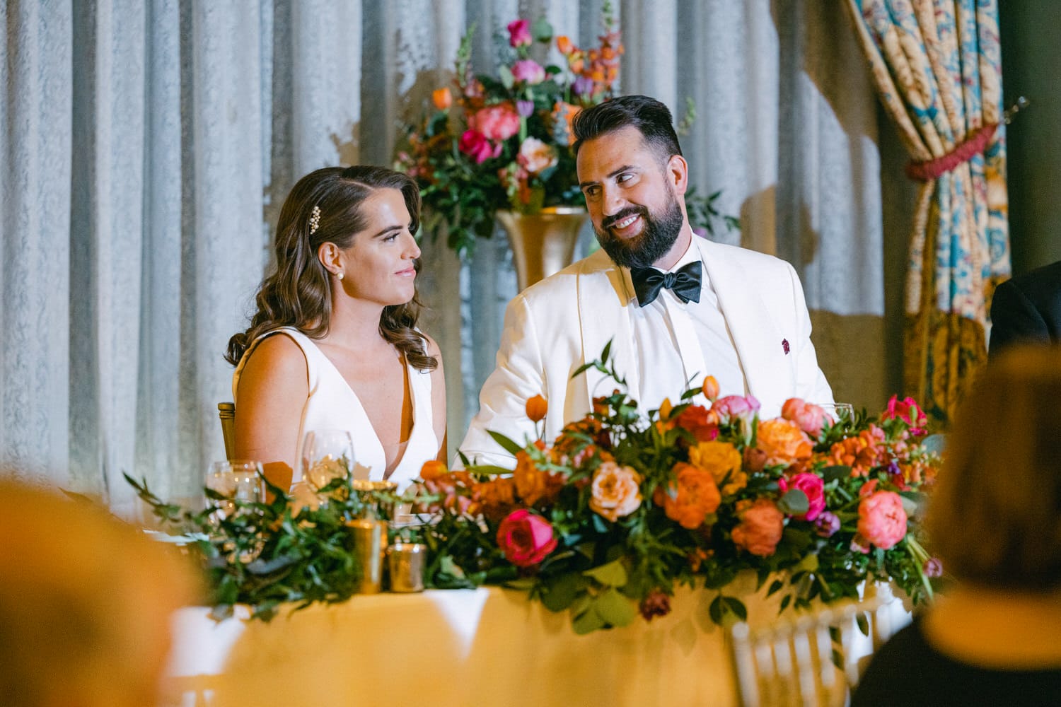 A beautifully decorated table with a couple smiling at each other, surrounded by vibrant floral arrangements during a wedding celebration.