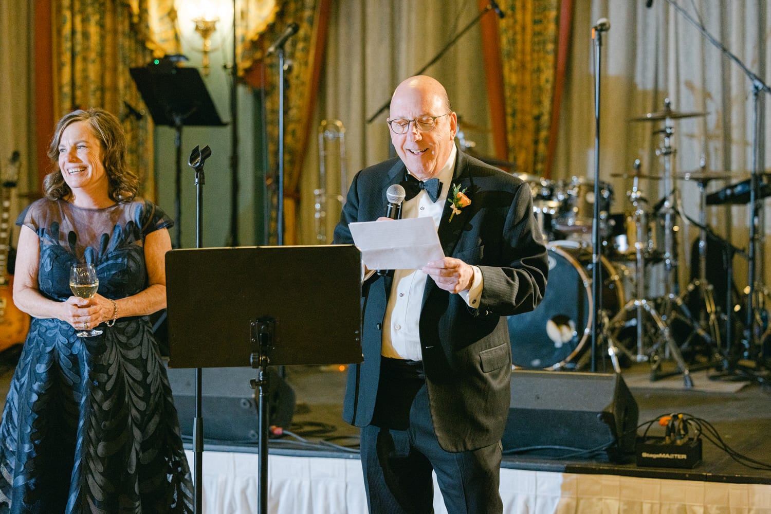 A man in a tuxedo delivers a speech while a woman in a dark dress smiles and holds a glass of wine, with a band setup in the background.