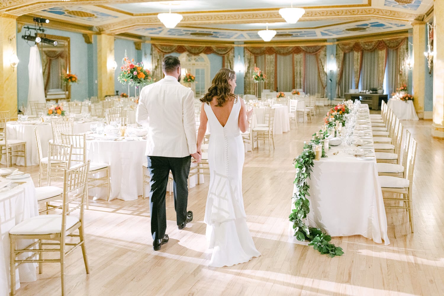 A couple walks hand-in-hand through a elegantly decorated wedding reception, featuring round tables adorned with floral centerpieces and a long table decorated with greenery.