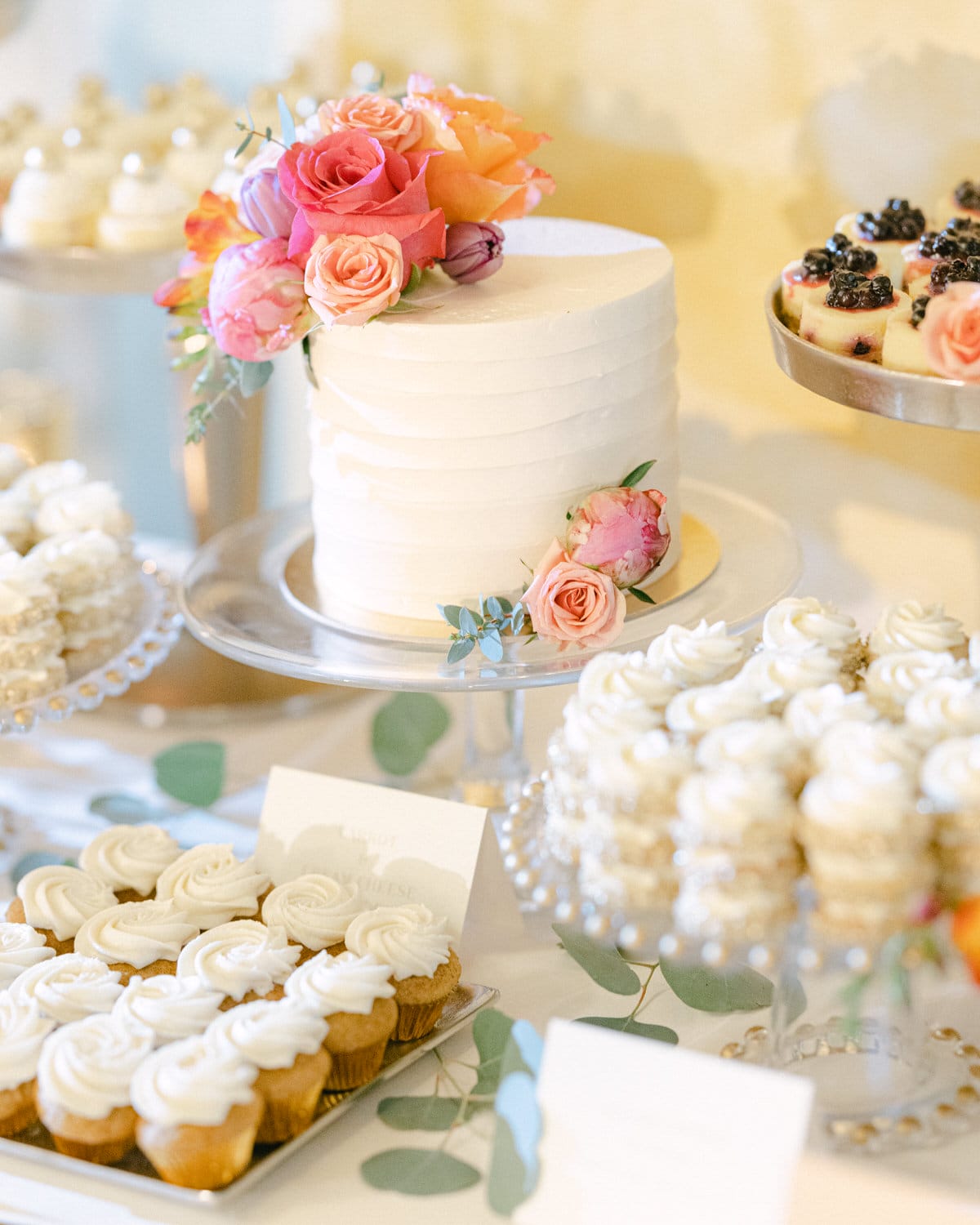 A beautifully arranged dessert table featuring a white cake adorned with colorful flowers, surrounded by elegant cupcakes and other treats.