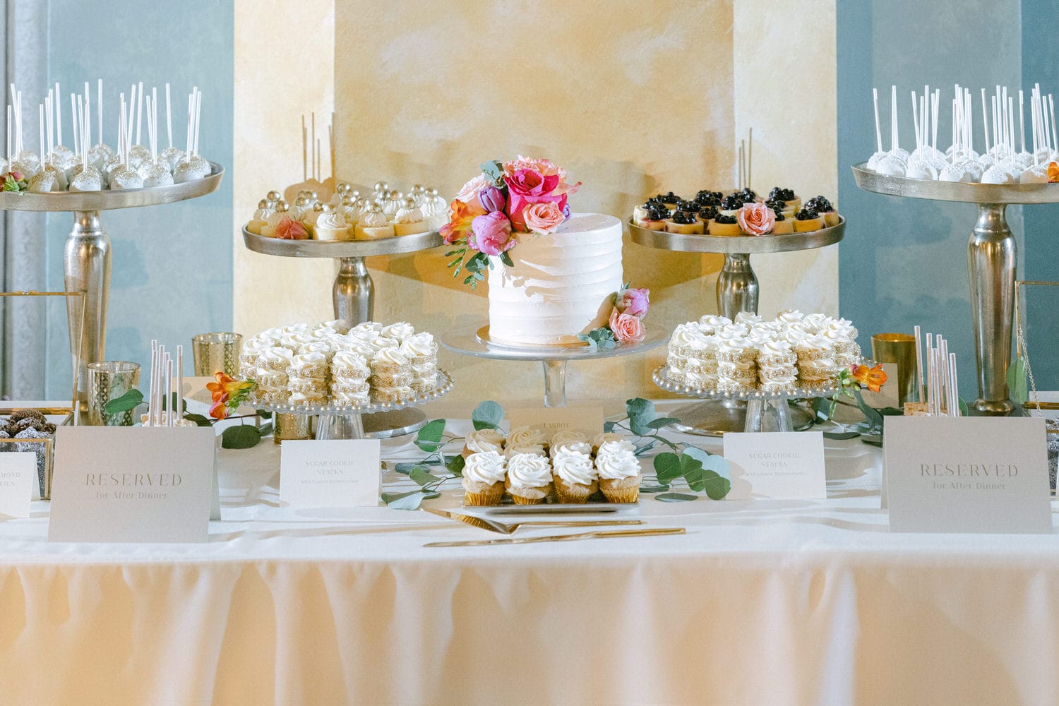 An elegant dessert table featuring a variety of beautifully decorated pastries, a floral-adorned cake, and a selection of reserved signs for guests.