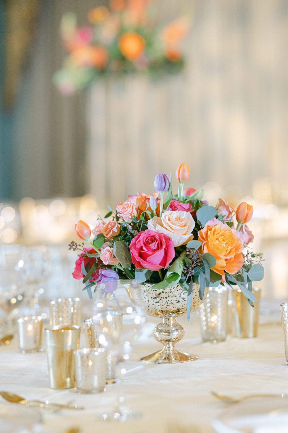 A vibrant arrangement of roses and tulips in a silver vase, surrounded by elegant table settings and soft bokeh in the background.