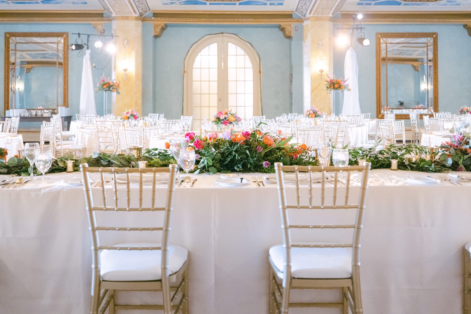 A beautifully arranged banquet space featuring tables adorned with floral centerpieces, elegant tableware, and golden chiavari chairs, all under a classic ornate ceiling.