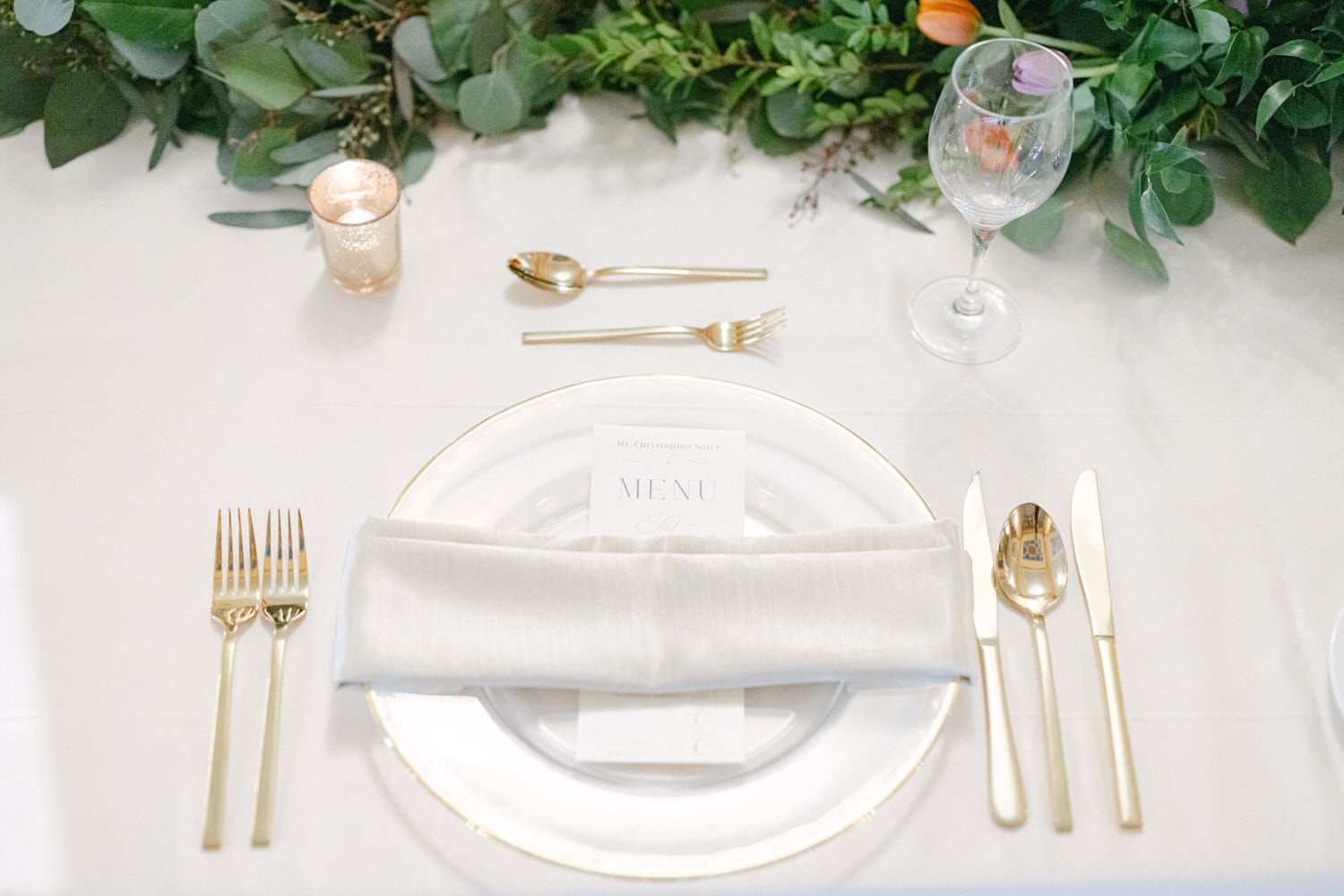 An elegantly arranged dining table featuring a gold-rimmed plate, utensils, a folded napkin, and a candle, complemented by greenery and floral accents.