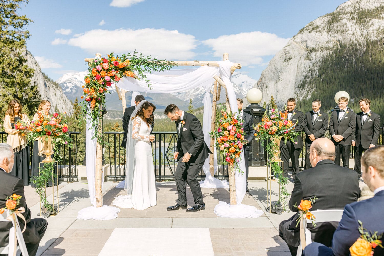 A couple exchanging vows under a floral arch with mountains in the background, surrounded by guests and a wedding party.