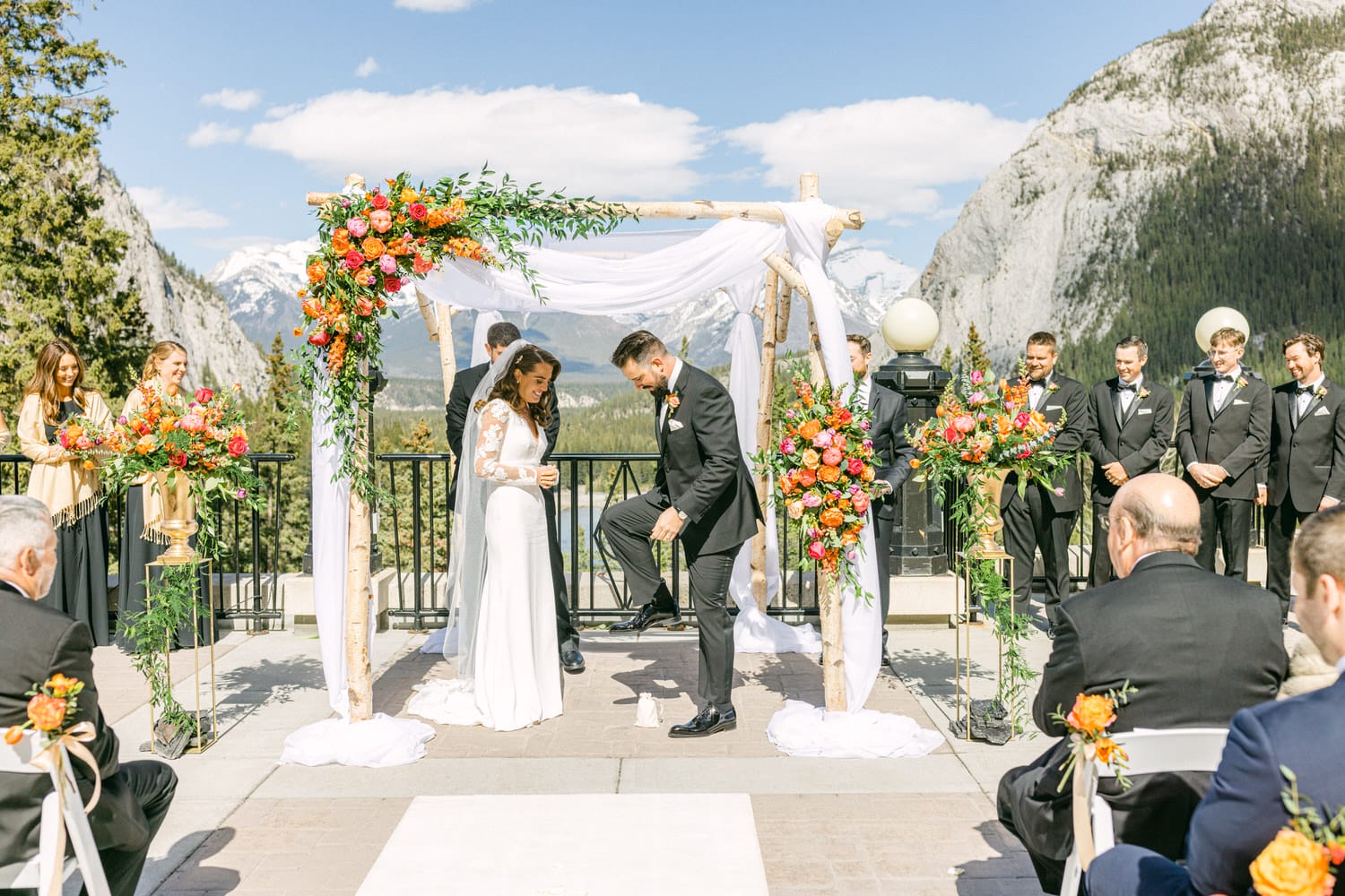 A joyful couple exchanges vows under a floral arch with mountains in the background, surrounded by family and friends.