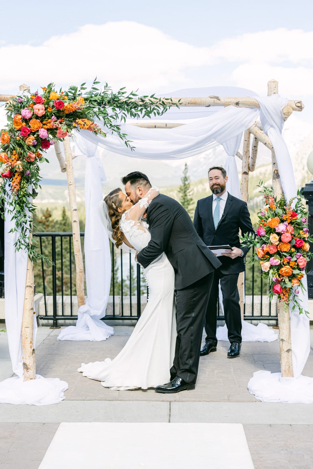 A couple shares a heartfelt kiss during their wedding ceremony under a floral arch, with a officiant standing nearby amidst a scenic background.