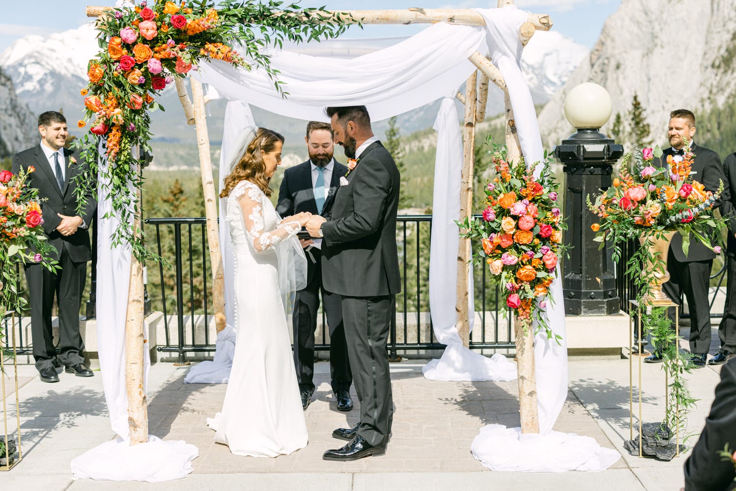 A couple exchanging vows under a floral-adorned arch, surrounded by wedding attendants in formal attire, with a stunning mountain landscape in the background.