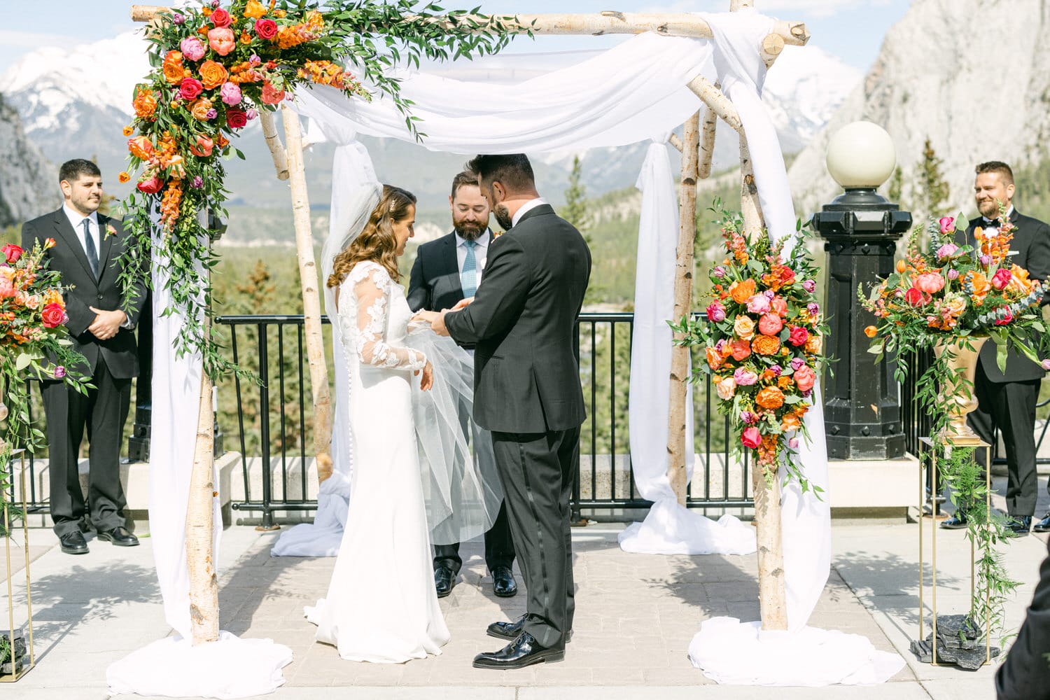 A couple exchanging vows under a floral arch with a mountain backdrop during an outdoor wedding ceremony.