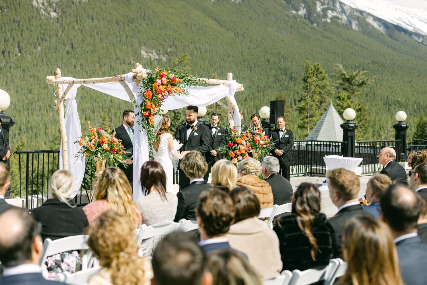 A couple exchanging vows under a floral arch at an outdoor wedding ceremony with guests seated in front, surrounded by lush green mountains.