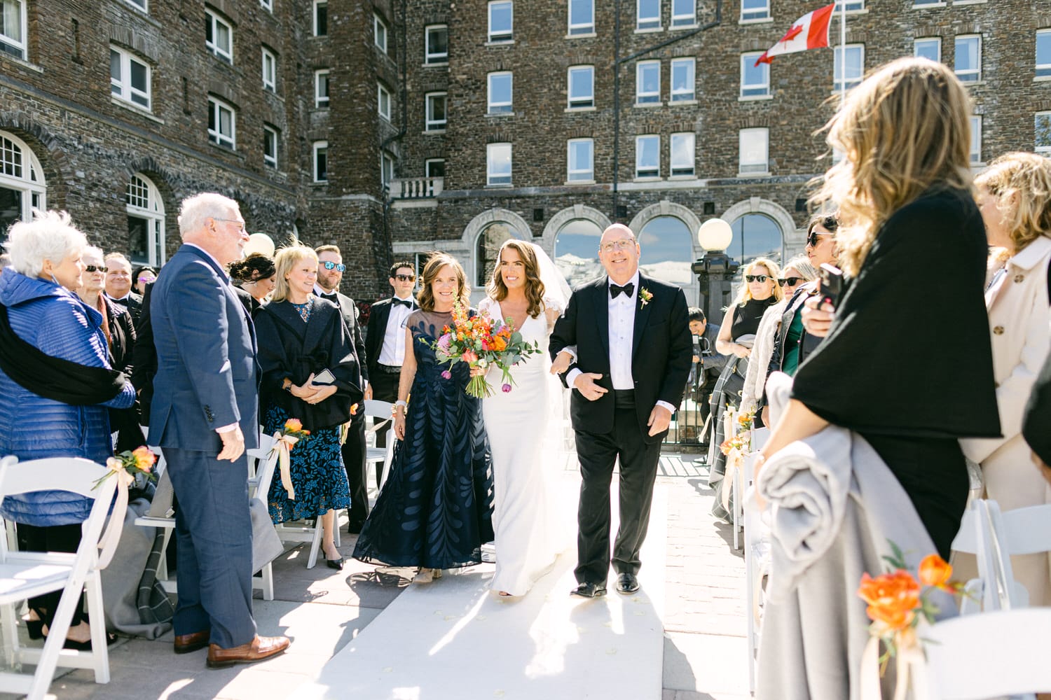 A bride walks down the aisle with her father, surrounded by guests in formal attire at an outdoor wedding venue. The bride smiles broadly, holding a colorful bouquet, while family and friends look on with joy.