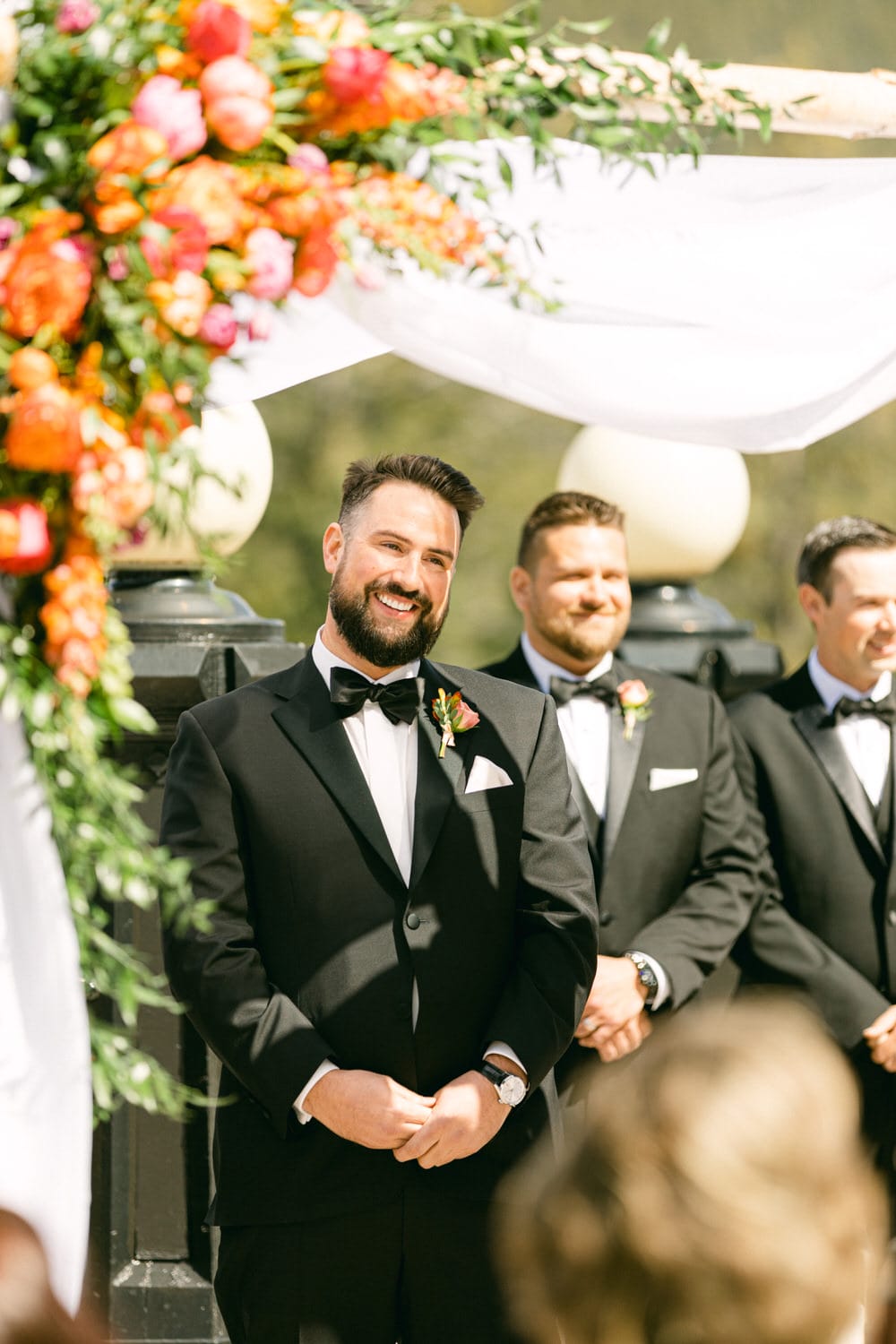 A groom smiling at the altar with his groomsmen beside him, adorned with vibrant floral arrangements above.