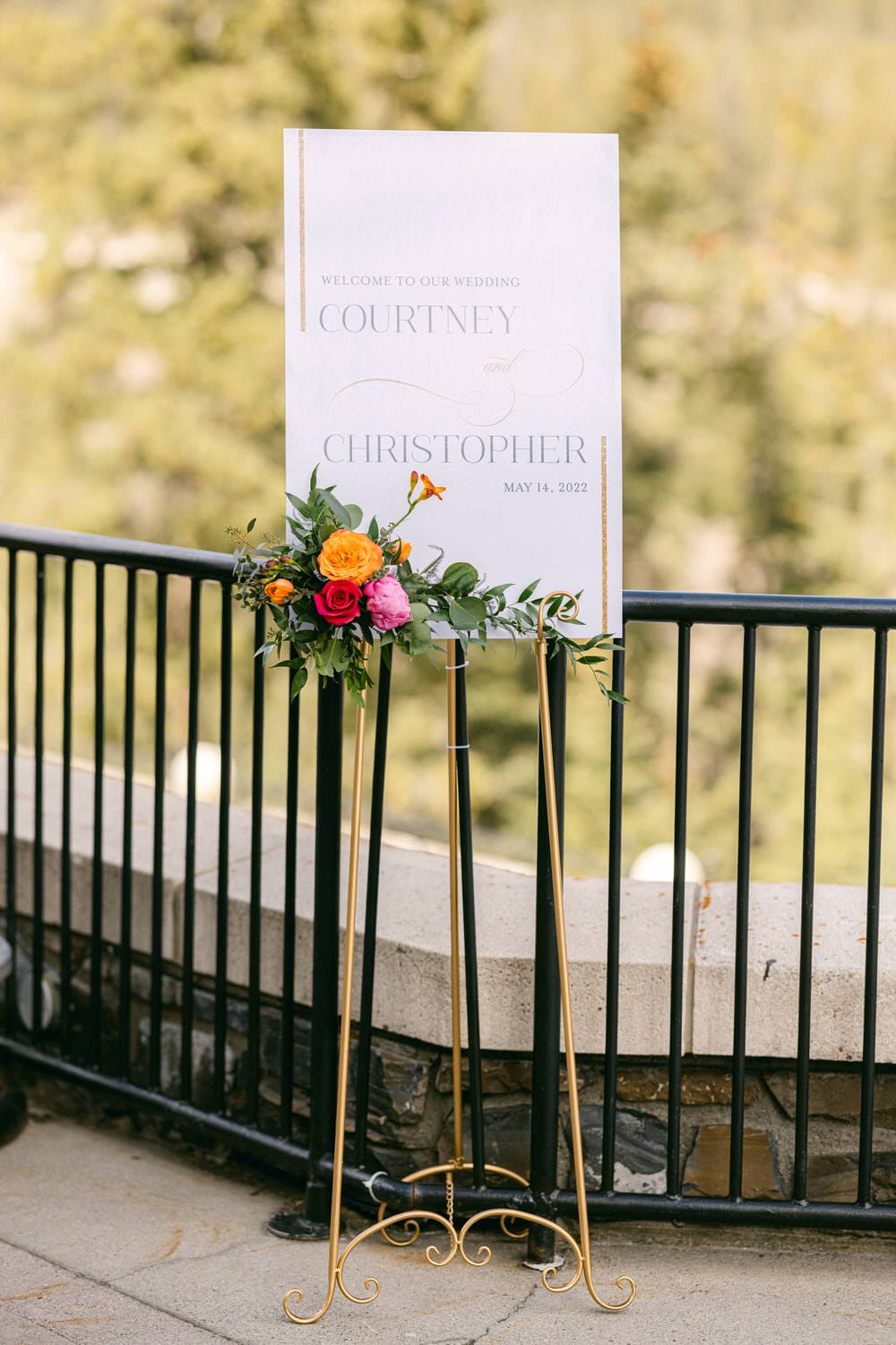 A beautiful wedding welcome sign displaying the names Courtney and Christopher, adorned with vibrant flowers, set outdoors against a lush green backdrop.