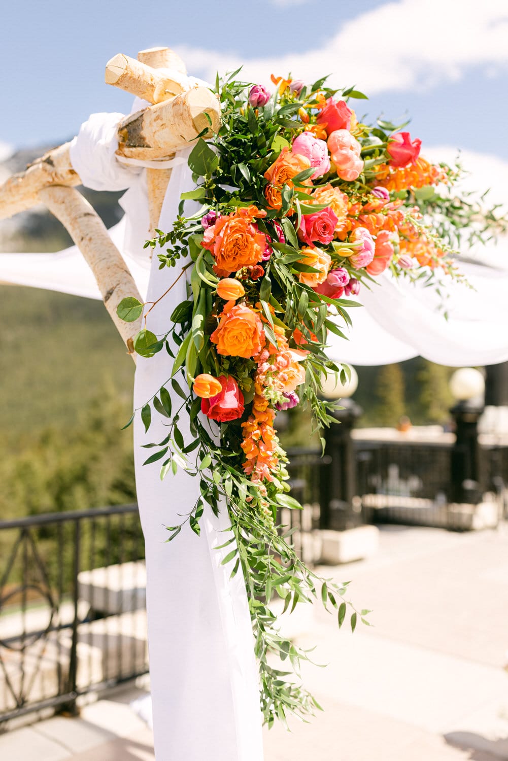 A beautifully arranged wedding arch adorned with an array of colorful flowers, including orange roses, pink tulips, and lush greenery, set against a scenic backdrop.