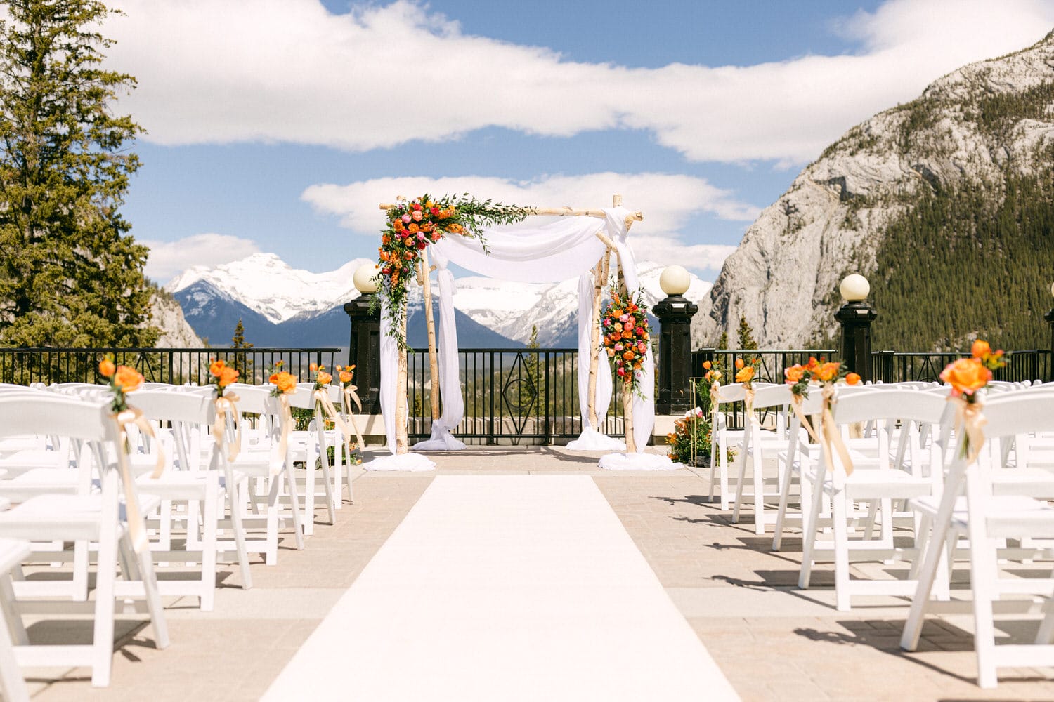 A beautifully arranged outdoor wedding altar adorned with flowers, white drapery, and chairs lined up for guests against a backdrop of mountains.