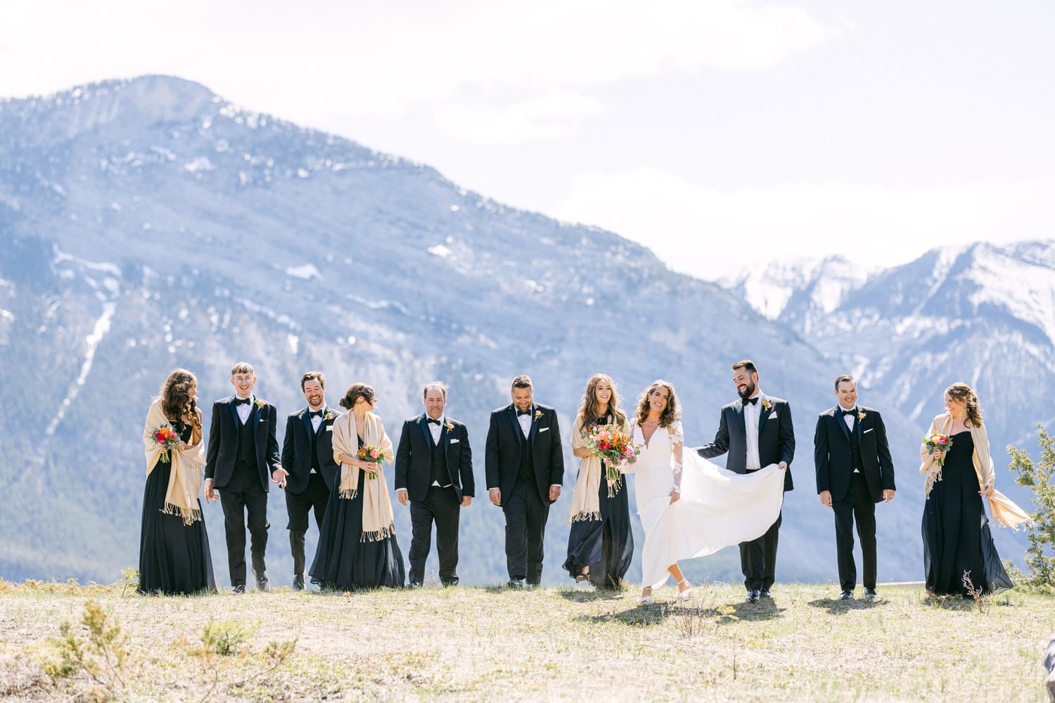 A joyful wedding party posing on a mountaintop, dressed in formal attire, with stunning mountains in the background.