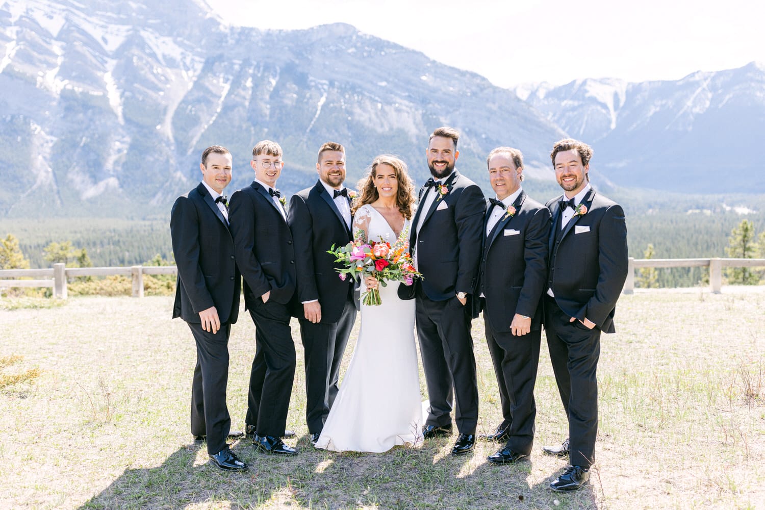 A joyful bride stands surrounded by her groom and six groomsmen, all dressed in formal black suits against a stunning mountain backdrop. The bride holds a colorful bouquet, exuding happiness on a sunny day.