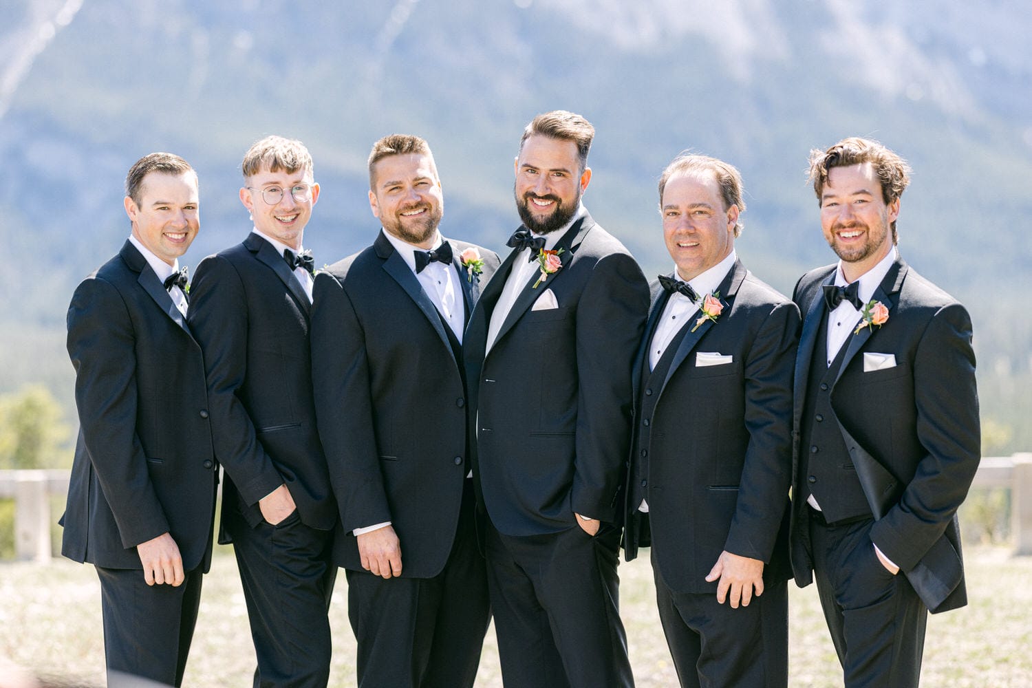 A group of six men dressed in tuxedos stands together outdoors, smiling against a scenic mountain backdrop.