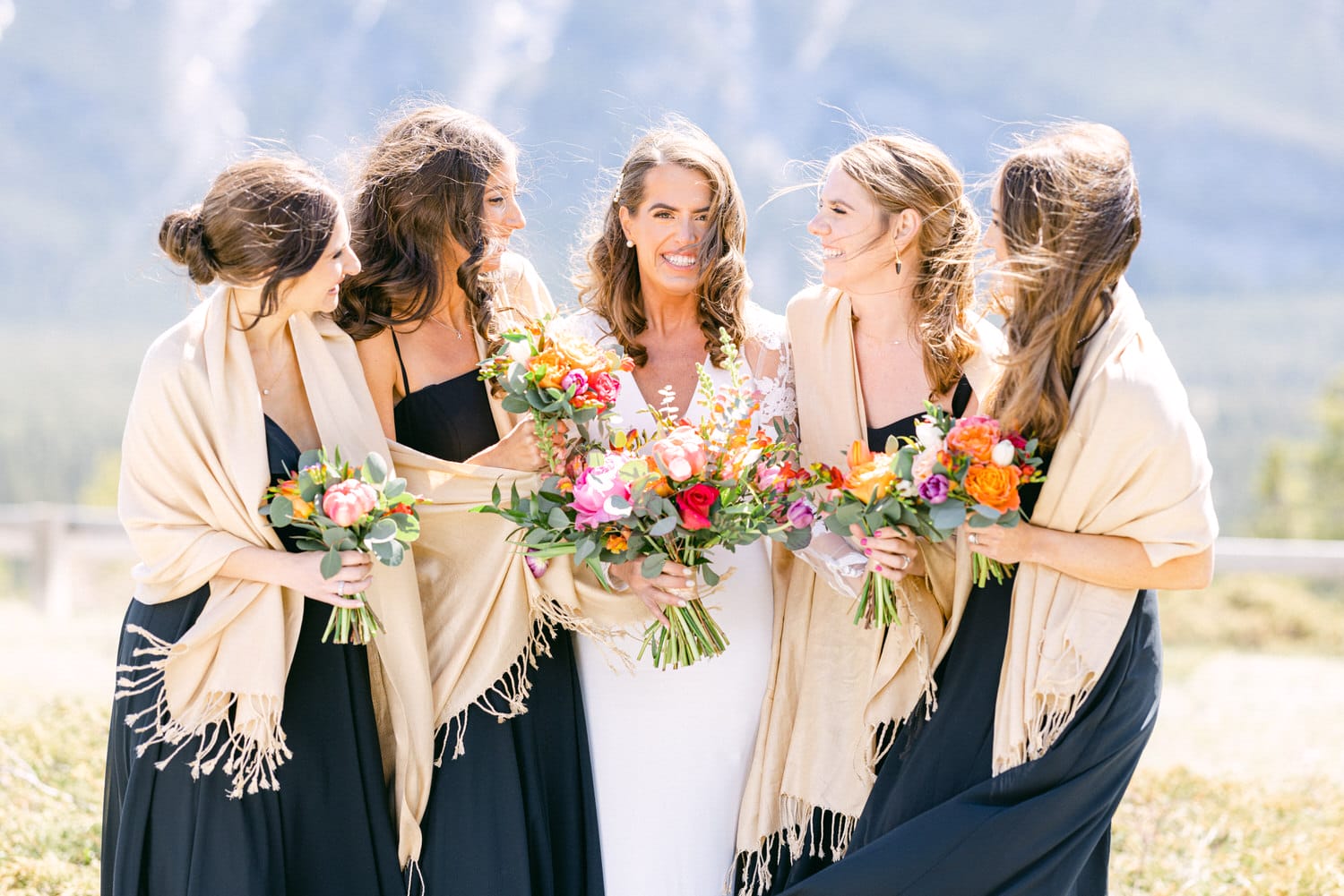 A joyful bride poses with her bridesmaids in elegant black dresses and shawls, surrounded by vibrant floral bouquets against a stunning natural backdrop.