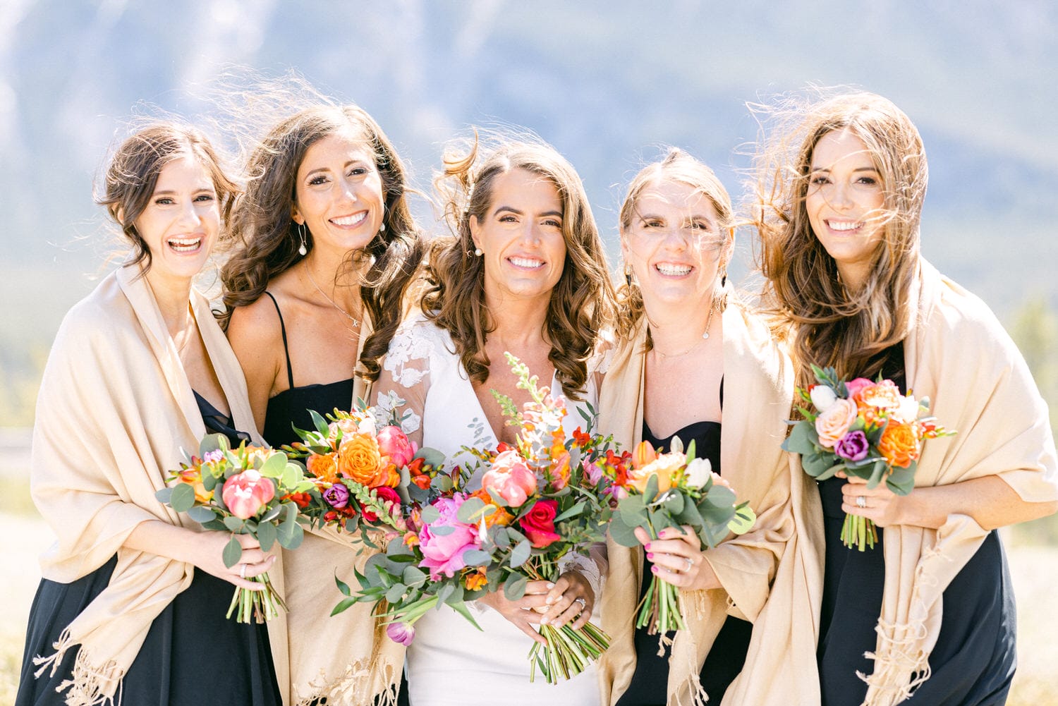 A cheerful bride and her four bridesmaids, dressed in elegant black dresses and shawls, holding vibrant floral arrangements against a scenic background.