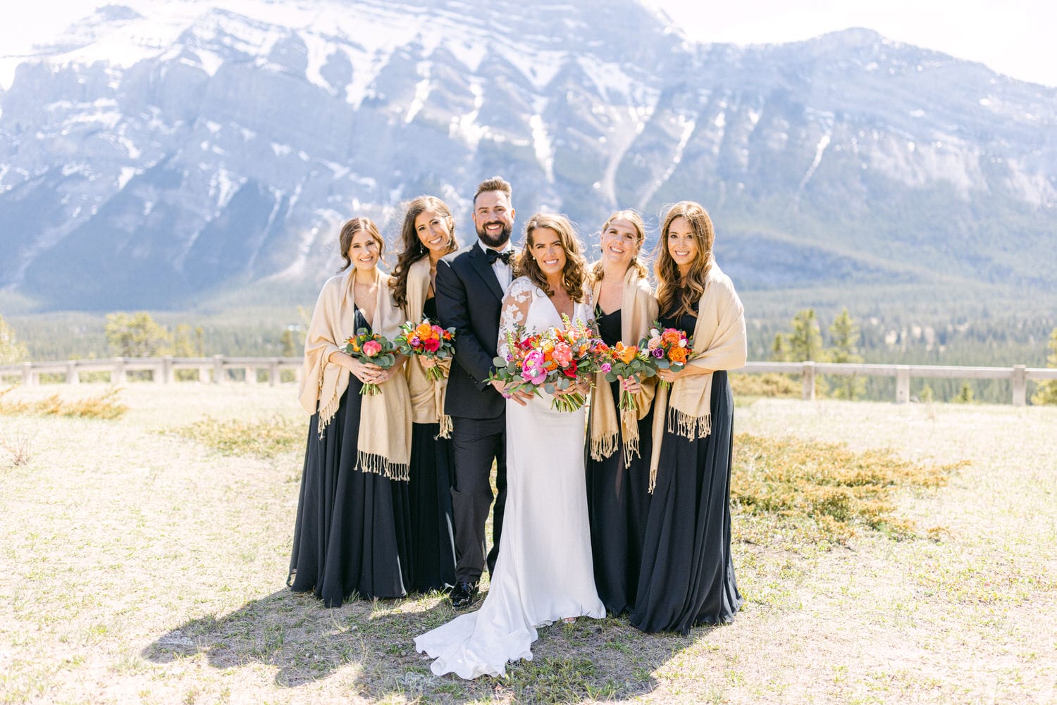 A bride and her bridesmaids pose together in front of majestic mountains, holding colorful bouquets and dressed in elegant attire.