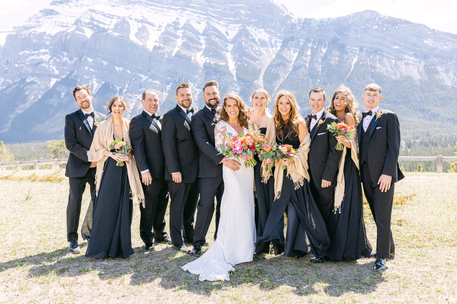 A joyful wedding party poses together in elegant attire with colorful bouquets, set against a stunning mountainous backdrop.