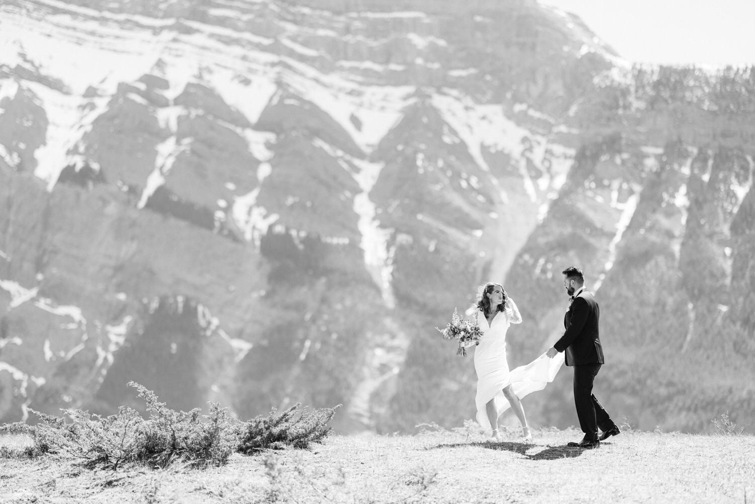 A joyful couple in formal attire celebrating their wedding against a stunning mountainous backdrop, with the bride holding a bouquet and her dress flowing in the wind.