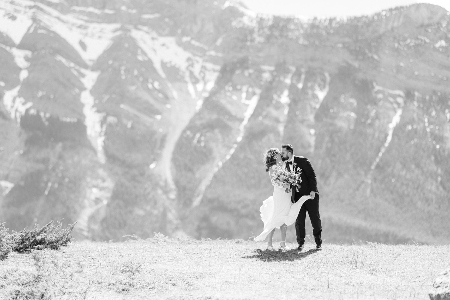 A couple shares a romantic kiss against a breathtaking mountain backdrop, capturing a moment of love and celebration on their wedding day.