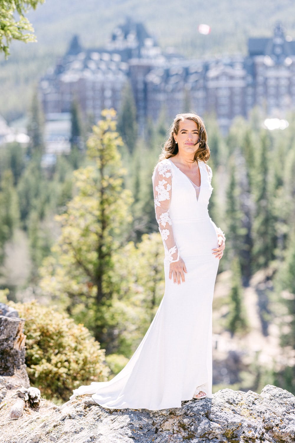 A bride in a white wedding dress poses gracefully on a rocky ledge surrounded by trees, with a grand hotel visible in the blurred background.