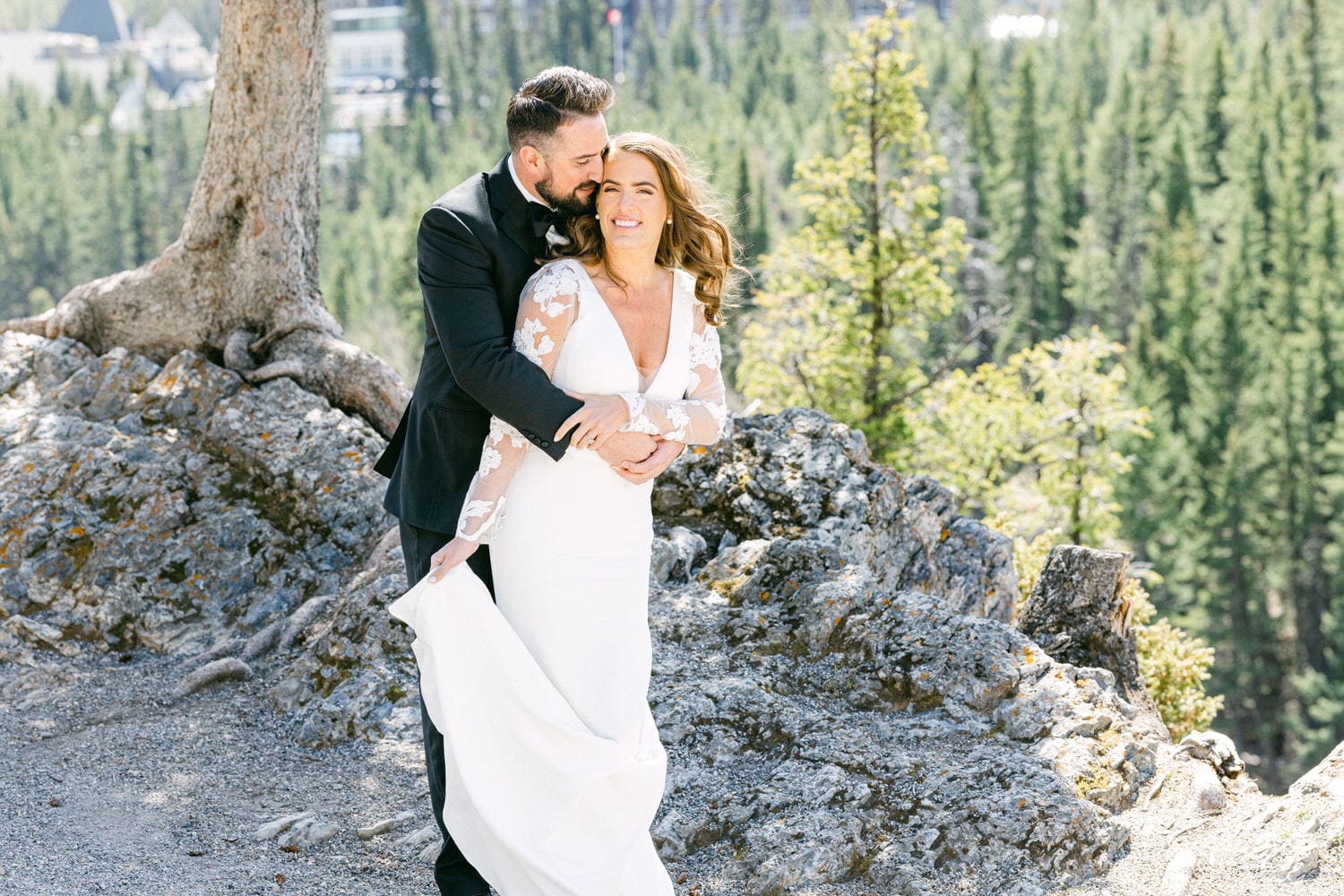 A joyful couple in formal wedding attire stands together outdoors, surrounded by lush green trees and rocky terrain, capturing a moment of love and happiness.