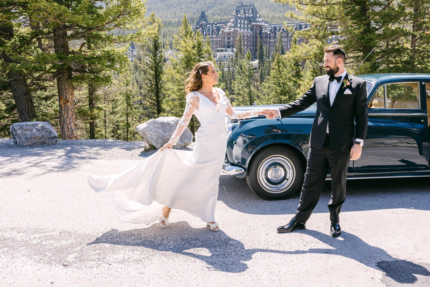 A bride and groom hold hands, joyfully posing near a vintage car, surrounded by a picturesque mountain backdrop and lush trees.