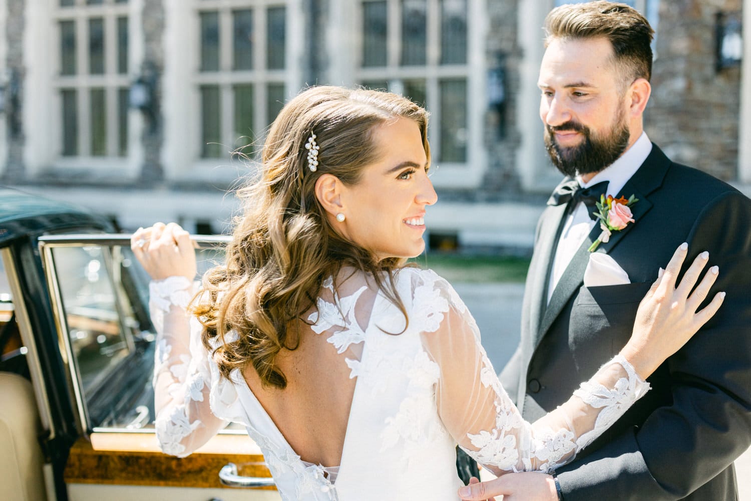 A bride in a stunning lace gown smiles as she holds the door of a classic car, while her groom looks on with admiration, set against a beautiful architectural backdrop.