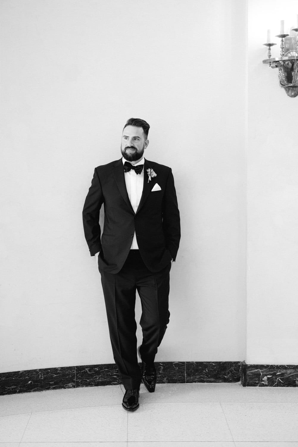 A well-dressed man in a tuxedo leans against a wall, exuding sophistication with a groom's boutonnière in his lapel.