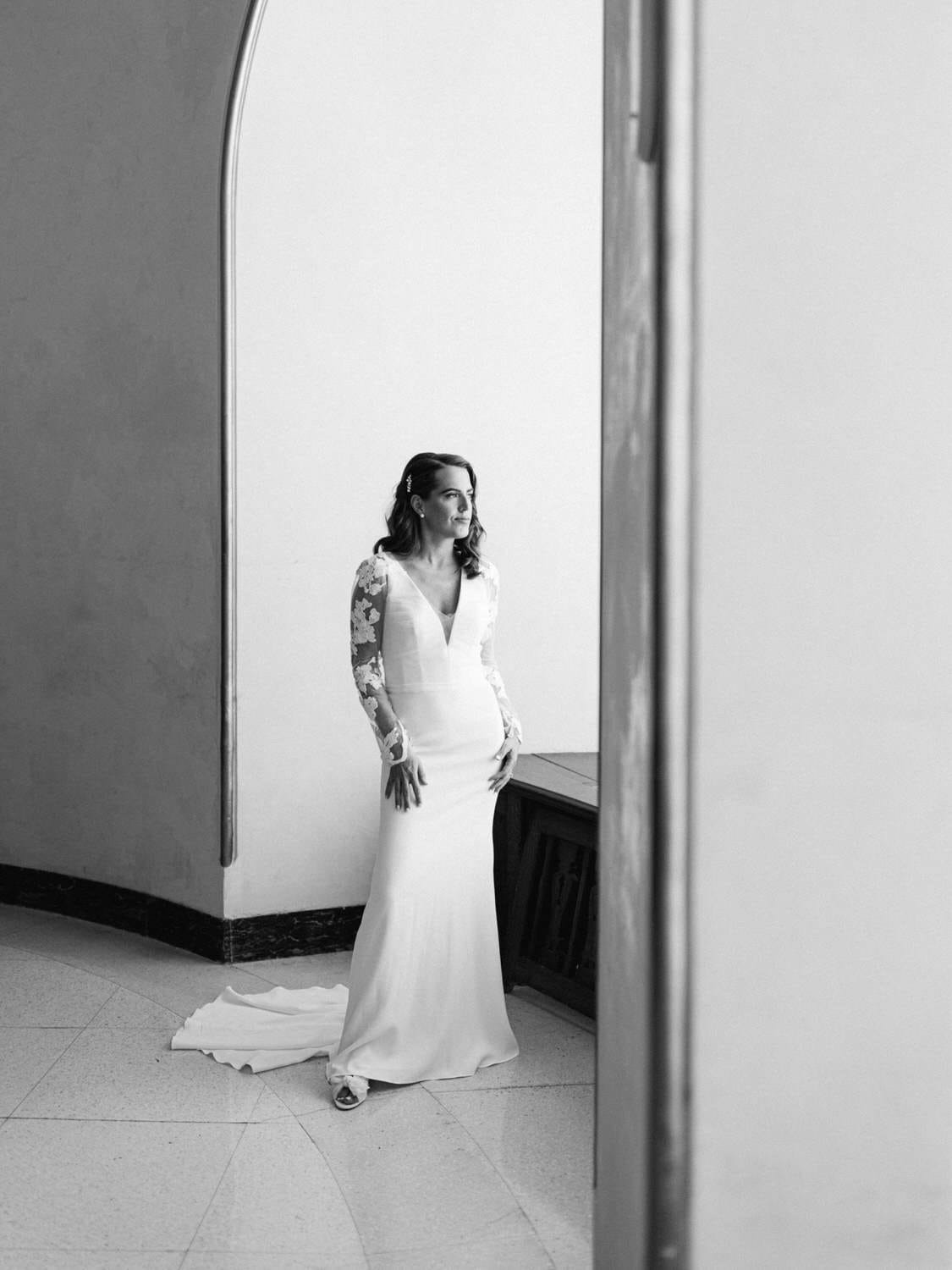 A poised bride stands gracefully in a fitted white gown with long lace sleeves, framed by a soft architectural archway.