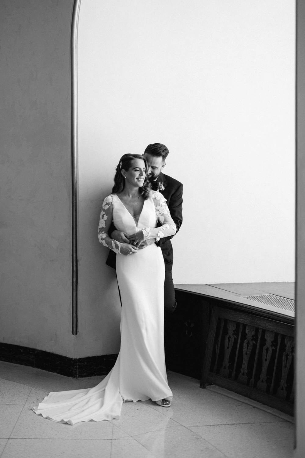 A couple embracing in a sophisticated setting, the bride in a sleek white gown with floral lace sleeves and the groom in a black suit, both sharing a joyful moment.