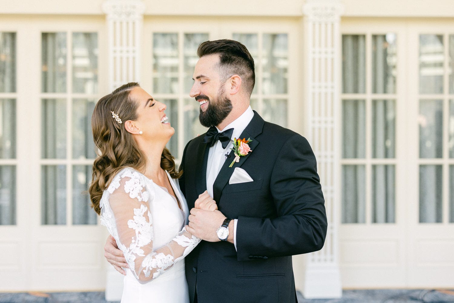 A bride and groom share a joyful moment, smiling and holding hands, set against a backdrop of elegant windows.