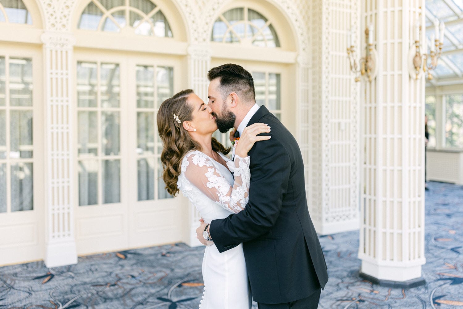 Bride and groom share a tender kiss in an elegant setting, surrounded by light and architectural details.