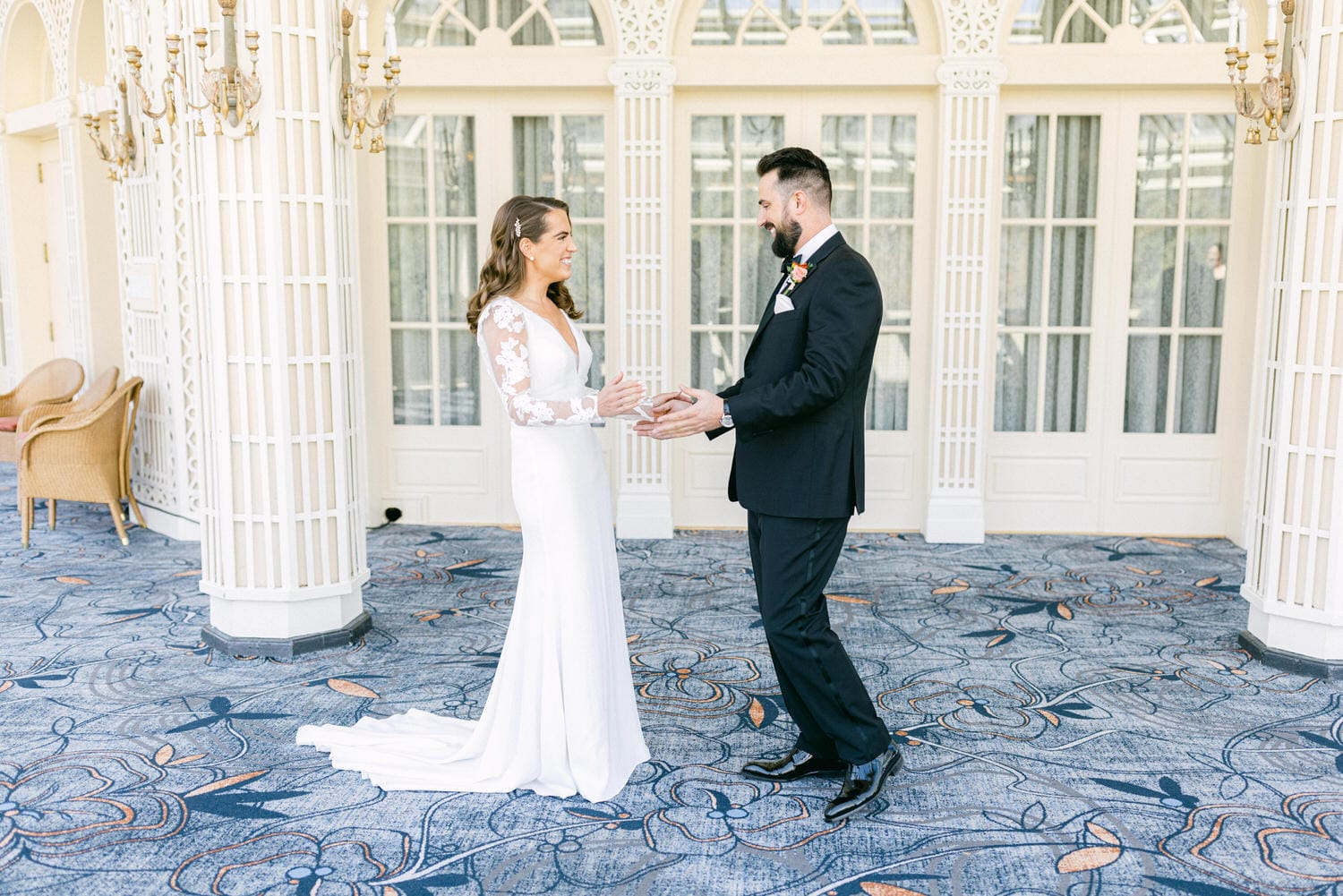 A bride in a sleek white gown and a groom in a black suit smile at each other in a beautifully decorated indoor space, showcasing their love and connection.