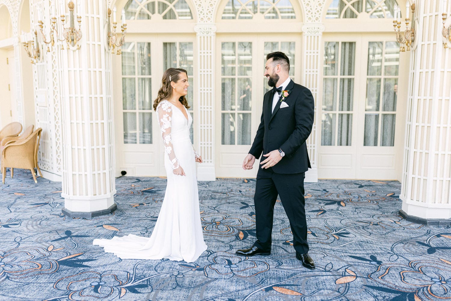 A bride in an elegant white gown and a groom in a black tuxedo share a joyful moment in a beautifully designed indoor space, with ornate columns and floral-patterned carpet.