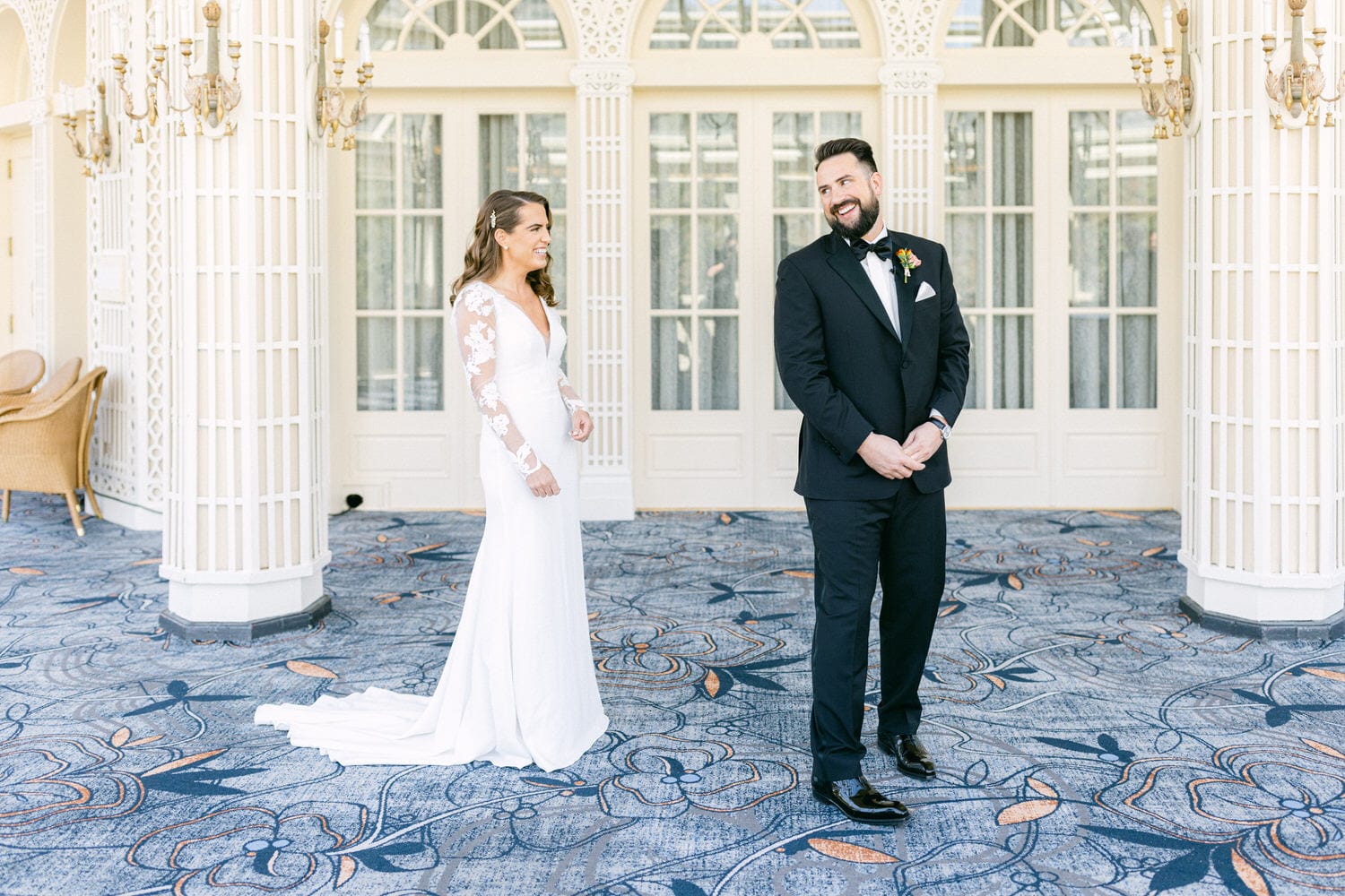 A bride in a long-sleeve wedding dress smiles at her groom, who is dressed in a tuxedo, against a backdrop of ornate architecture and floral-patterned carpet.