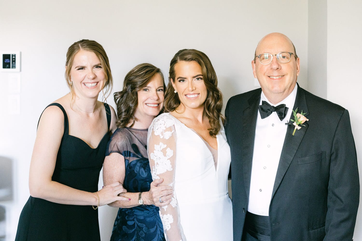 A smiling bride stands with her mother, father, and sister, all dressed elegantly, showcasing love and joy in a well-lit setting.