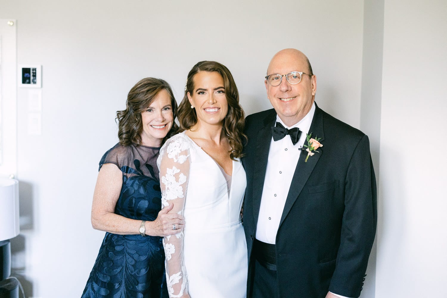 A happy bride in a white gown poses with her smiling mother and father, dressed in elegant attire, in a well-lit indoor setting.