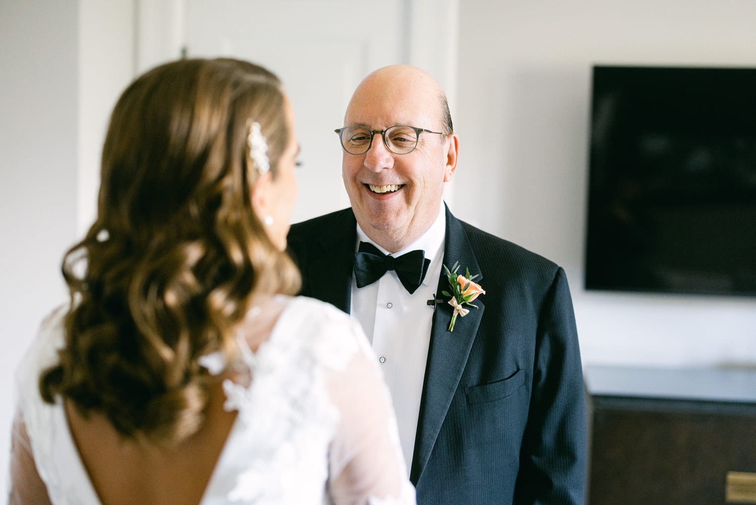 A joyful daughter in a wedding dress smiles at her father, who is wearing a tuxedo and glasses, both sharing a heartfelt moment.