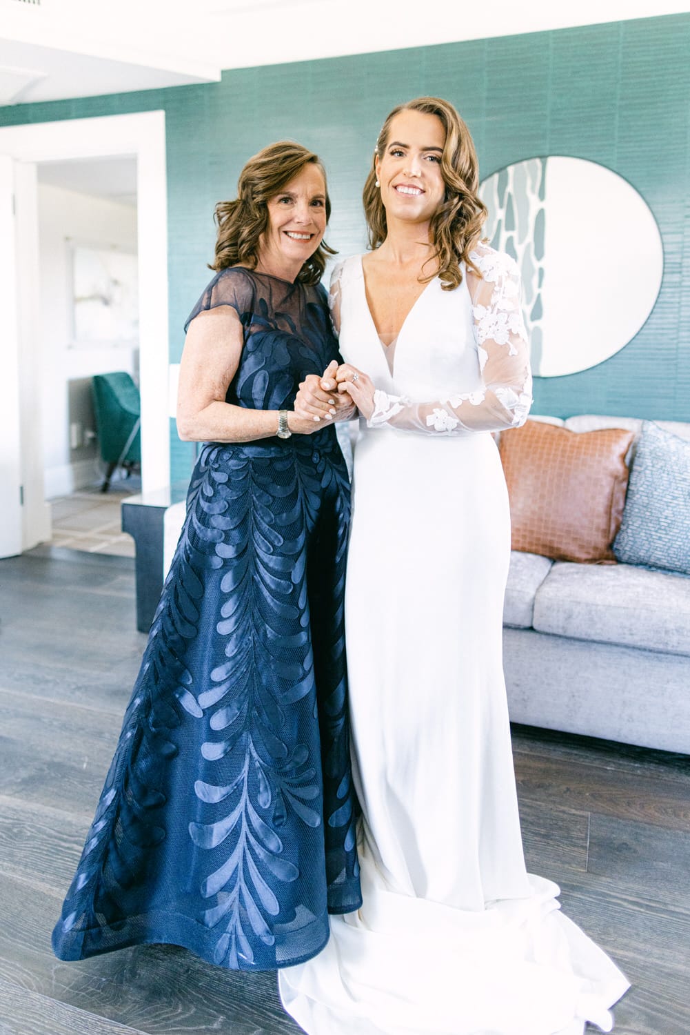 A joyful bride in a white gown poses with her mother, both smiling and holding hands, set against a stylish interior backdrop.