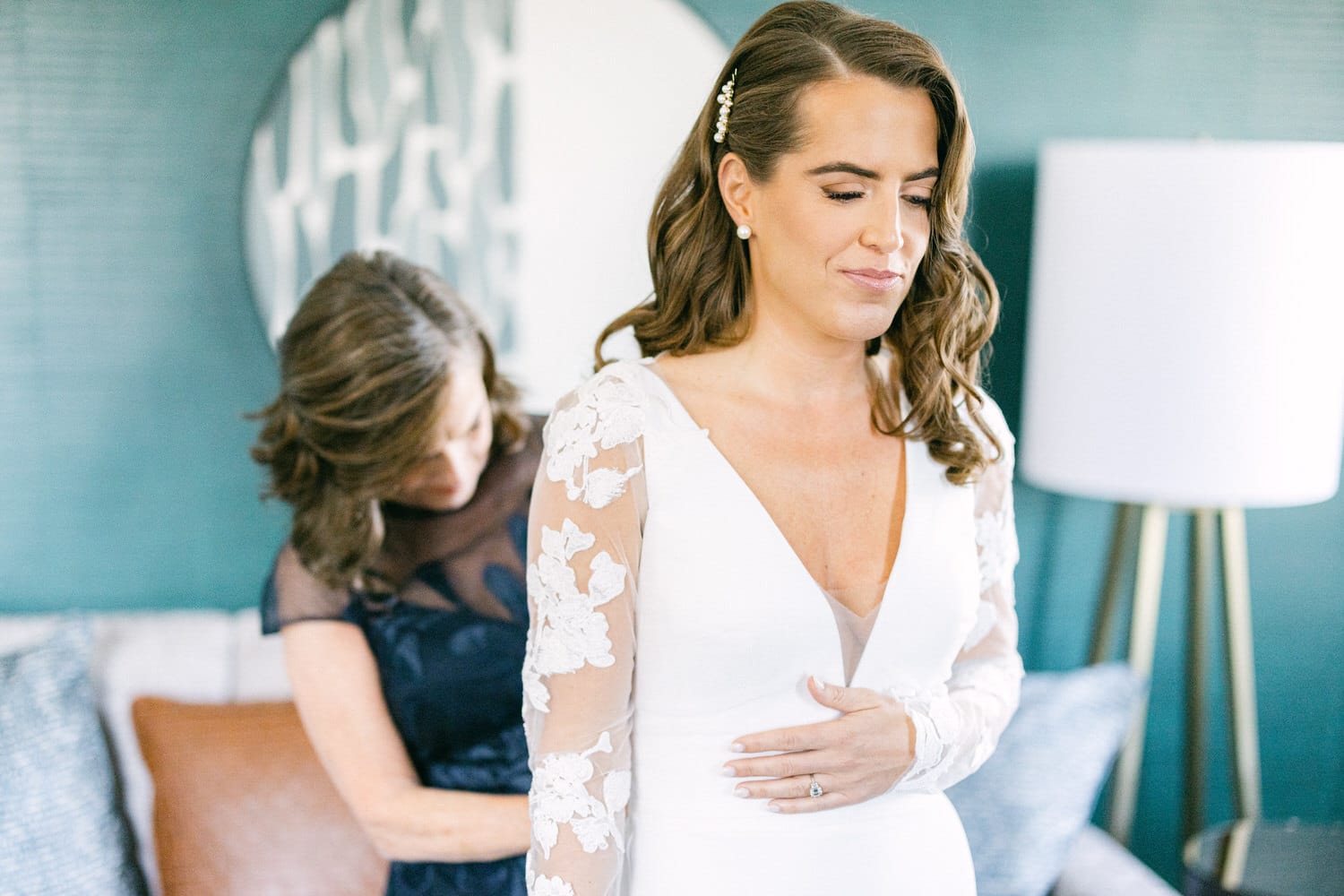 A bride in a white dress with floral lace sleeves stands with a serene expression while a woman adjusts her gown from behind, set in a stylishly decorated room.