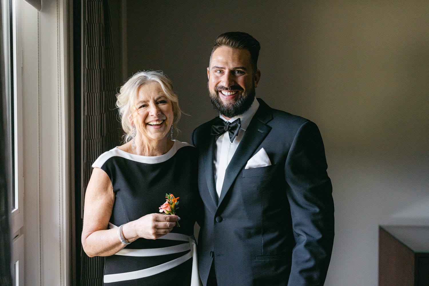 A smiling mother and her son posing together, dressed elegantly for a special occasion, with soft natural light illuminating the scene.