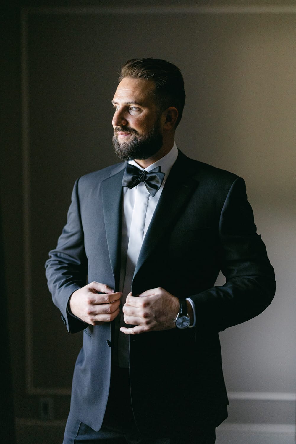 A well-groomed man in a tuxedo adjusts his bow tie while standing in soft, warm light, exuding elegance and confidence.