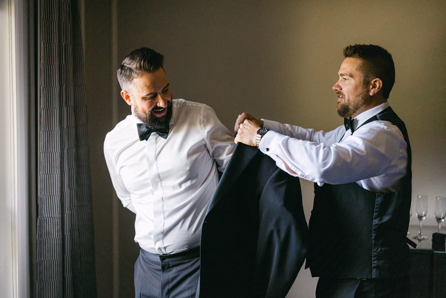 Two men help each other get dressed in formal attire, with one adjusting a suit jacket on the other in a well-lit room.