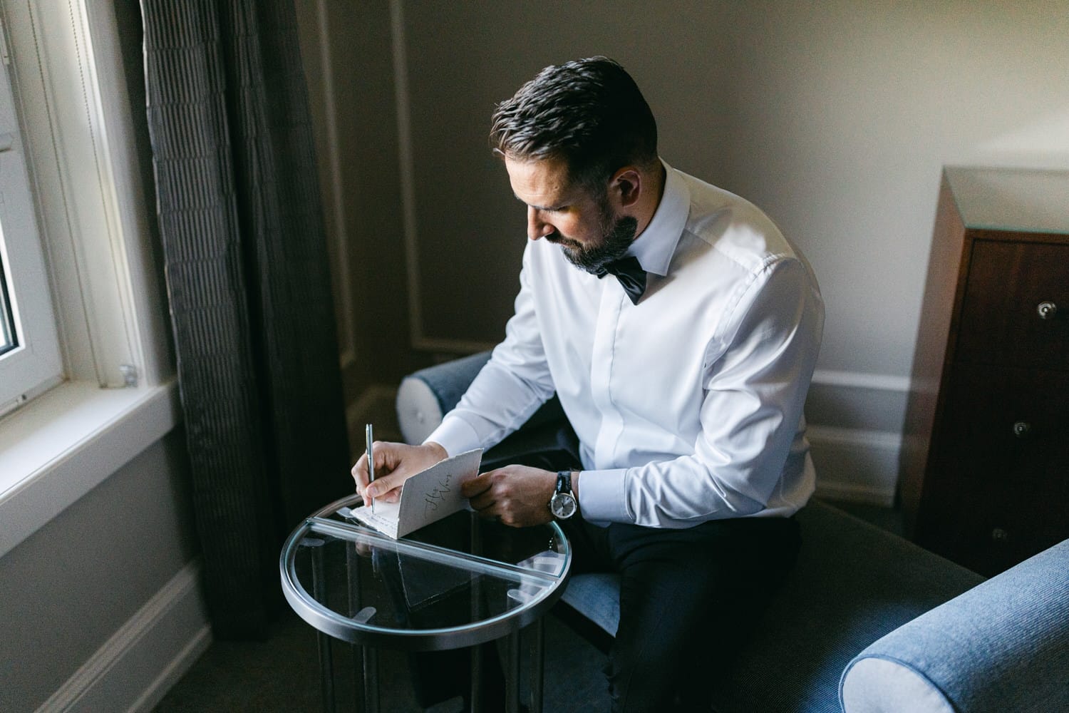 A groom writes a personal note while sitting on a chair, dressed in a white shirt and bow tie, with a modern clear table beside him.